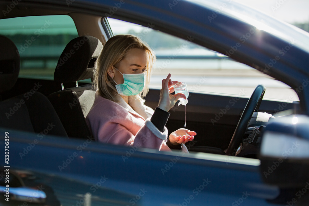 Woman in protective mask sitting in a car on road, using hand sanitizer. Safe traveling. - obrazy, fototapety, plakaty 