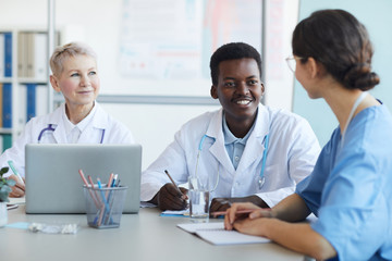 Wall Mural - Multi ethnic group of doctors sitting at table, focus on young African-American man in committee meeting at medical council