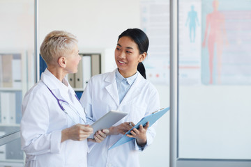 Wall Mural - Waist up portrait of young female doctor talking to supervisor and smiling cheerfully while standing in medical office interior, copy space