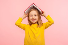 Portrait Of Happy Lazy Little Girl Covering Head With Book And Smiling To Camera, Disobedient Child Having Fun, Fooling Around Instead Studying Lesson. Indoor Studio Shot Isolated On Pink Background