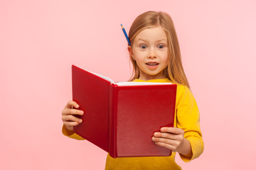 Shocking facts. Astonished smart little girl with pencil behind ear reading big book with surprised expression, amazed by story, learning encyclopedia. indoor studio shot isolated on pink background