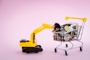 The excavator loader model with pile of coins in the shopping cart on pink background for saving money, investment, business and finance concept