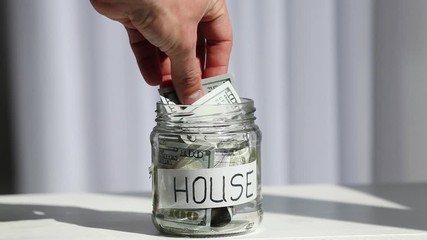 Wall Mural - Male hand puts 100 dollars in a glass jar with the text house. Concept of saving money.