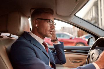 Talking with client. Handsome businessman in full suit adjusting headphones while driving a car