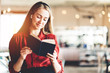 © Alex from the Rock - Portrait of young woman reading book wearing wireless earphones, standing in bright open space room
