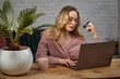 © nazarovsergey - Female in pink cardigan, glasses. Working on laptop and showing white plastic card. Sitting at wooden table with pink cup, palm in pot on it. Close up