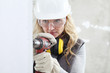 © amedeoemaja - woman contruction worker using cordless drill driver making a hole in wall, builder with safety hard hat, hearing protection headphones, gloves and protective glasses, close up portrait