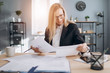 © Kuz Production - Portrait of concentrated woman in eyeglasses sitting at table in modern office with documents and doing paper work. Mature lady with blond hair in formal clothing skillfully doing her job.