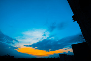 Wall Mural - Atmospheric blue cloudy sky behind silhouettes of city buildings. Cobalt and orange background of sunrise with dense clouds and bright yellow sunny light for copy space. Cyan heaven above clouds.