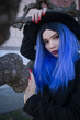 © Ruslan - Street Fashion young attractive girl with blue hair, dark lipstick on her lips in a black hat and dark clothes on a background of unusual trees with twisted branches and urban locations