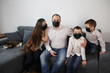 © kittyfly - young family, a man, a woman and two sons are sitting in a room on a gray sofa in black viral masks, the concept is a cozy home, love, happy parenthood during a viral epidemic