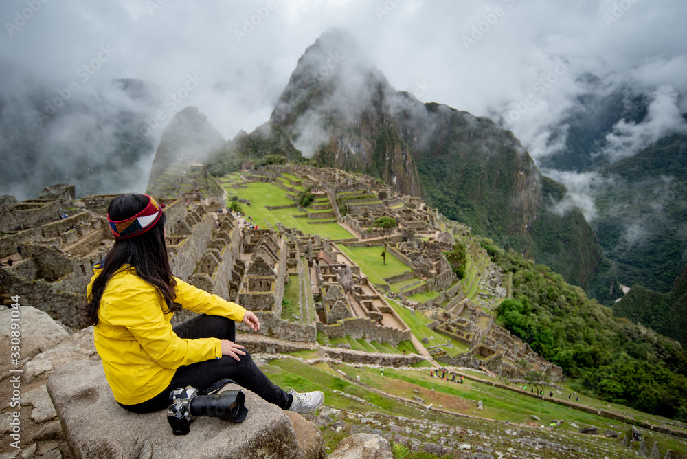 Young Asian woman traveler looking at Machu Picchu, one of seven wonders and famous tourist attraction in Cusco Region of Peru. This majestic place has known as 'Lost City of the Incas' - obrazy, fototapety, plakaty 