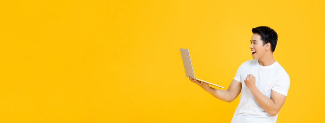 Happy young Asian man carrying laptop computer and raising his fist doing yes gesture isolated on yellow banner background with copy space