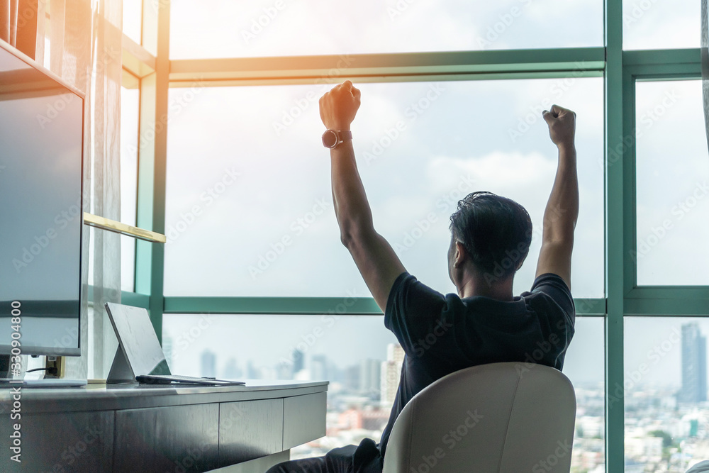 Business achievement concept with happy businessman relaxing in office or hotel room, resting and raising fists with ambition looking forward to city building urban scene through glass window - obrazy, fototapety, plakaty 