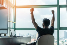 Business Achievement Concept With Happy Businessman Relaxing In Office Or Hotel Room, Resting And Raising Fists With Ambition Looking Forward To City Building Urban Scene Through Glass Window