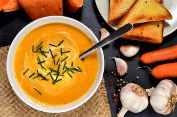 Top view to bowl of sweet potato cream soup with fresh chive and spoon, pepper, carrot, garlic and roasted toast bread on slate slab.