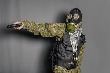 A soldier in camouflage uniform, body armor, in a gas mask with a hood on his head, holds a gun in his hands. Cosplay man, stalker. Studio photo on a gray background.