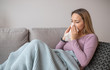 © makyzz - Sick woman blowing nose, sitting under the blanket. Sick woman with seasonal infections, flu, allergy lying in bed. Sick woman covered with a blanket lying in bed with high fever and a flu, resting.