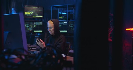 Caucasian woman cyber policewoman with blond short hair sitting in dark room with monitors and tapping on smartphone. Female cyberpolice browsing online on mobile phone. Hacker scrolling on cellphone.