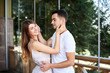 © Natalia - Young couple in love hugging by wooden railing, glass window and green trees on terrace. Blond woman, wearing stripy short overall and brunette man in white t-shirt and blue shorts on romantic date.