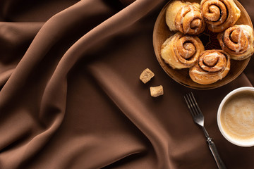 Wall Mural - top view of fresh homemade cinnamon rolls on silk brown cloth with brown sugar, fork and coffee