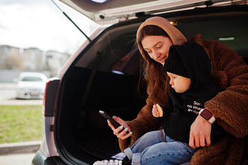 Young mother and child sitting in the trunk of a car and looking at mobile phone. Safety driving concept.