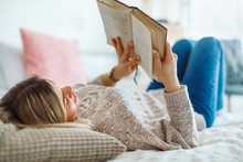 Young Woman Lies On Her Back In Bed Reading A Book