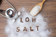 White Granulated Natural Sea Salt In Wooden Scoop And Words" Low Salt " Letters Written In Salt Grains And Medical Stethoscope On Wood Table Background. Unhealthy Food Concept. Top View. Flat Lay.