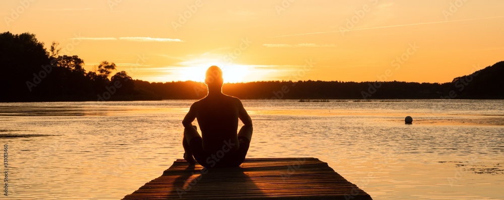 man sitting alone on a jetty by the lake at sunset in summer - obrazy, fototapety, plakaty 