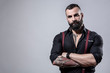 © alex.pin - Ragazzo barbuto con Tatuaggi in tutto il corpo, vestito con camicia nera e bretelle rosse,, incorcia le braccia,  isolato su sfondo grigio