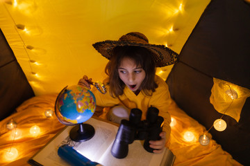 Wall Mural - young female child using magnifying glass to explore earth globe in a home made livingroom tent with
