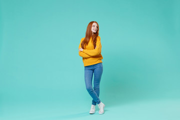 Wall Mural - Pretty young redhead woman girl in yellow sweater posing isolated on blue turquoise wall background studio portrait. People lifestyle concept. Mock up copy space. Holding hands crossed, looking aside.