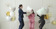 Stock video of three happy and cheerful girlfriends wearing pyjamas jumping on bed and fighting with pillows. White room is decorated with white and gold balloons.