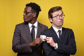 Young man in suit giving bribe to business partner on gyellow background.