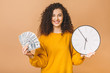 © denis_vermenko - Portrait of a cheerful young woman holding money banknotes and clock, celebrating isolated over beige background.