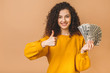 © denis_vermenko - Portrait of a cheerful young curly woman holding money banknotes and celebrating isolated over beige background. Thumbs up.
