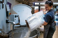Woman Wearing Apron Standing In An Artisan Bakery, Pouring Flour Into Industrial Mixer.