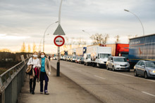 Defocused View Of Two Women Walking With Protective Surgical Masks Border Between Germany And France With Large Queue Of Cars Crisis Against The Novel Coronavirus.