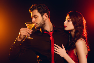 Wall Mural - Handsome man drinking cocktail near elegant girlfriend on black background with lighting