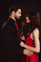 Wall Mural - Side view of handsome man hugging elegance girlfriend with rose isolated on black