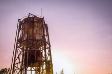 Low Angle Shot Of Two Old Water Tank Towers Against A Setting Sun With Purple Sky