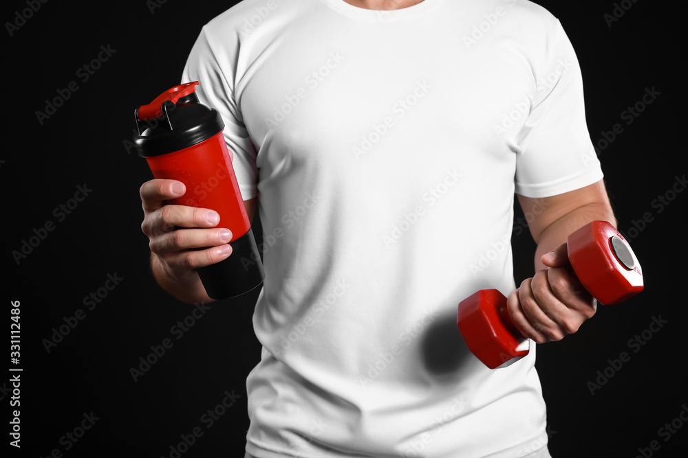 Sporty man with protein shake and dumbbell on dark background