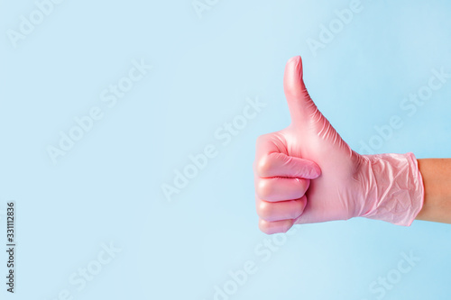 Closeup of female hand in pink cosmetic medical hygiene pharmacy gloves showing thumbs up sign against pastel blue background, copy space, minimal concept