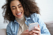 © insta_photos - Cheerful afro american millennial woman holds smart phone watching social media stories video. Happy young mixed race teen girl using funny mobile app laughing, playing game, chatting on cell at home.