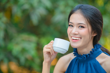 Close up beautiful asian woman with blue dress fresh morning drinking hot coffee  on sunny day; Happy young business woman  holding white coffee cup in hand over park background.