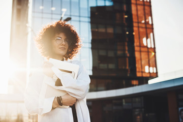 Lovely caucasian freelancer with curly hair and eyeglasses is posing confidently outside while holding a laptop