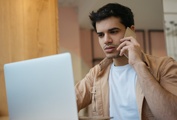 Indian man talking on mobile phone, working from home. Portrait of pensive asian businessman using laptop computer, planning project, communication in modern office 