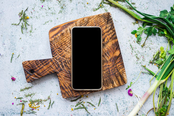 Empty black phone screen on a wooden board at kitchen table with greens.  Menu order or online food buy concept service.