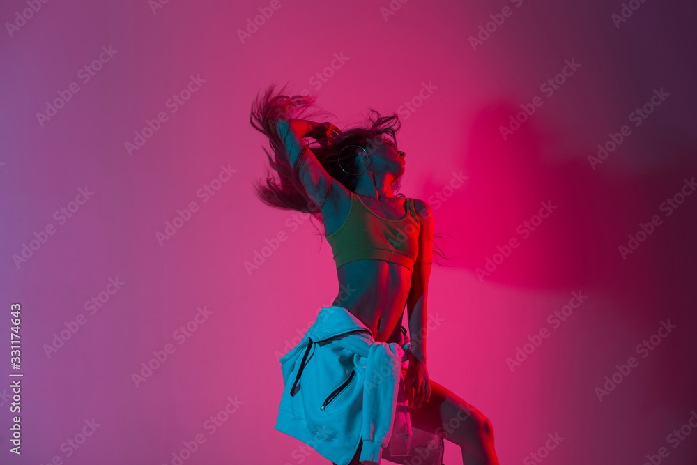 Fashionable young woman model in stylish youth clothes dancing in the studio with amazing bright neon pink color in disco style. Modern slim sexy girl dancer posing indoors with multi-colored light. - obrazy, fototapety, plakaty 