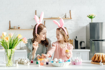 happy child and mother with bunny ears near chicken eggs, decorative rabbits, easter bread and tulips